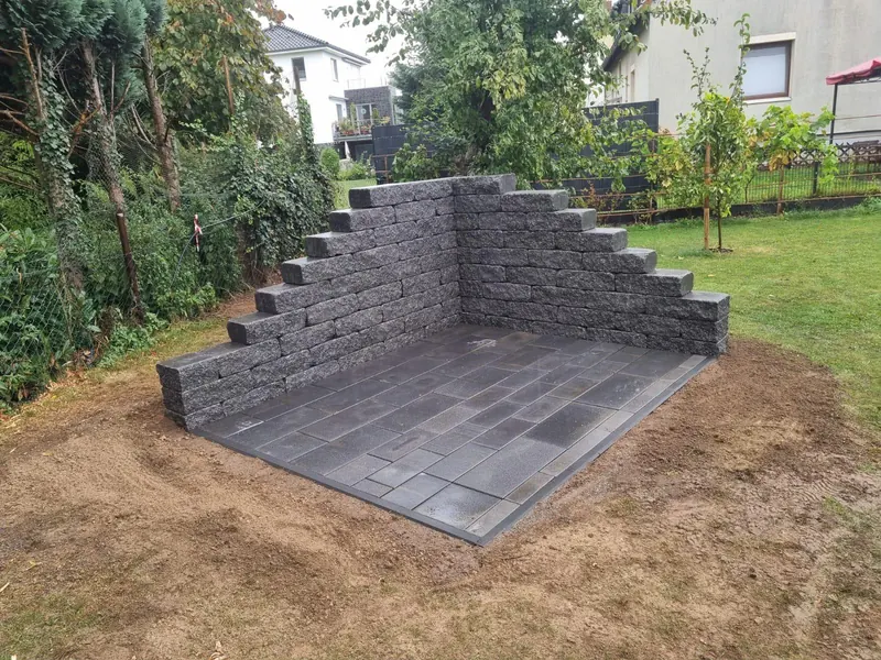 Gestufte Natursteinoptik-Mauer aus anthrazitfarbenen Mauersteinen mit Plattenbelag